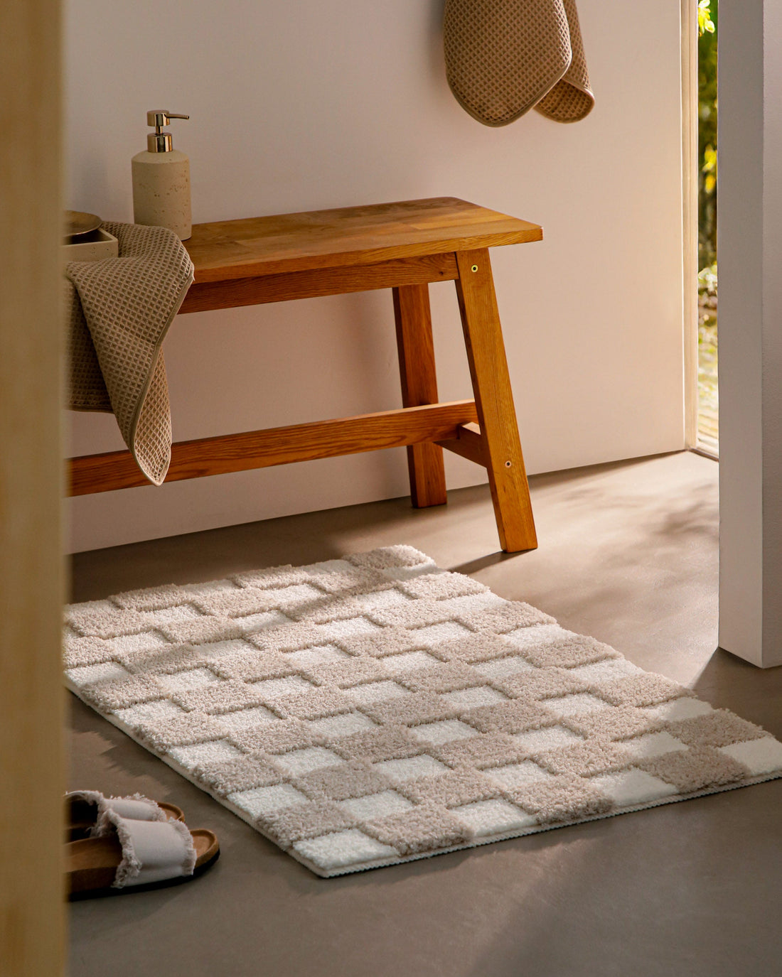 Badteppich von BathKing by WECONhome Bobbi Beige im Badezimmer neben Holzbank auf hellem Boden liegend, rechteckige Matte mit Karomuster