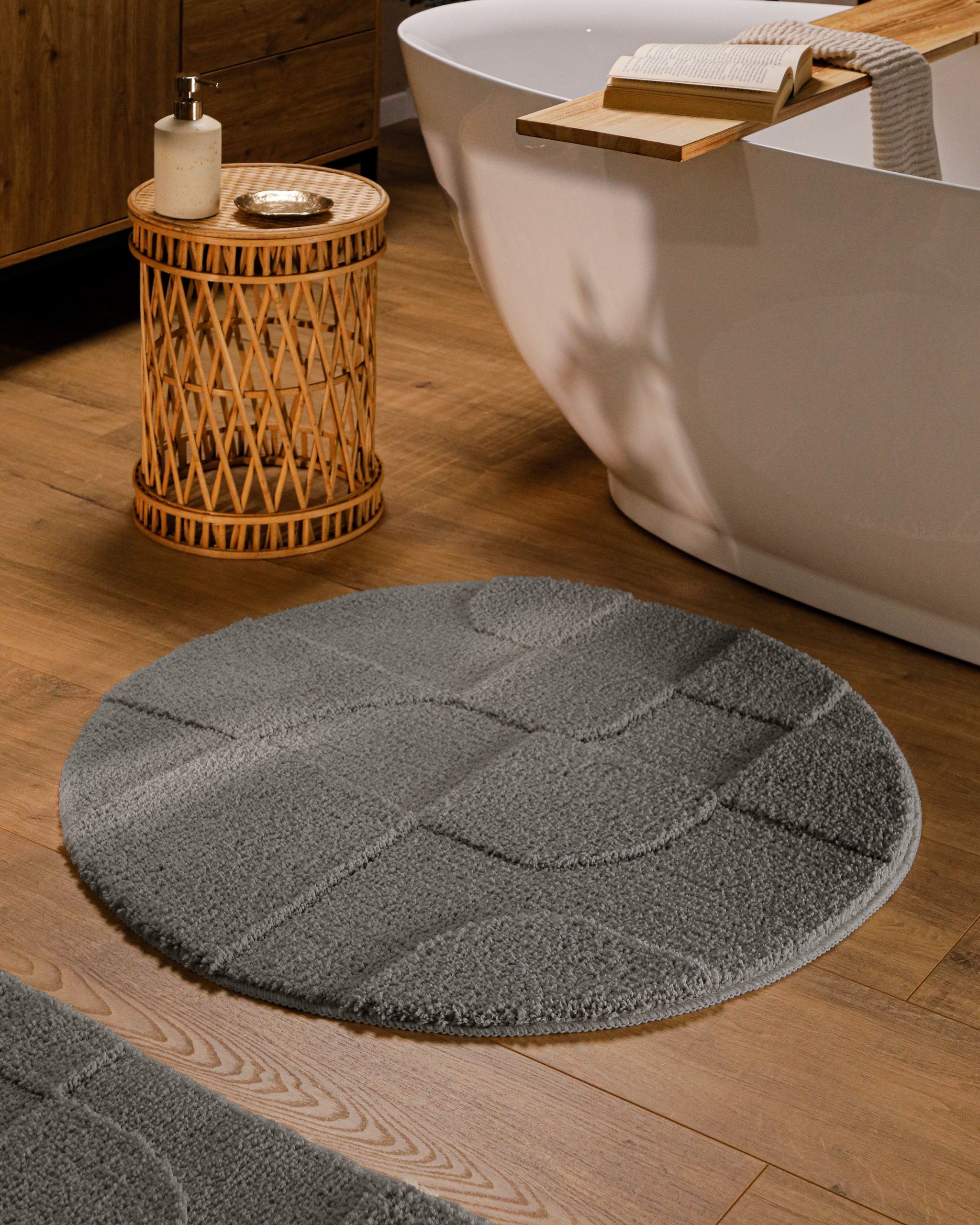 Runder Badteppich von BathKing by WECONhome Davie Basalt Grau im Badezimmer vor Badewanne auf Holzboden, strukturiertes Linienrelief im Flor sichtbar