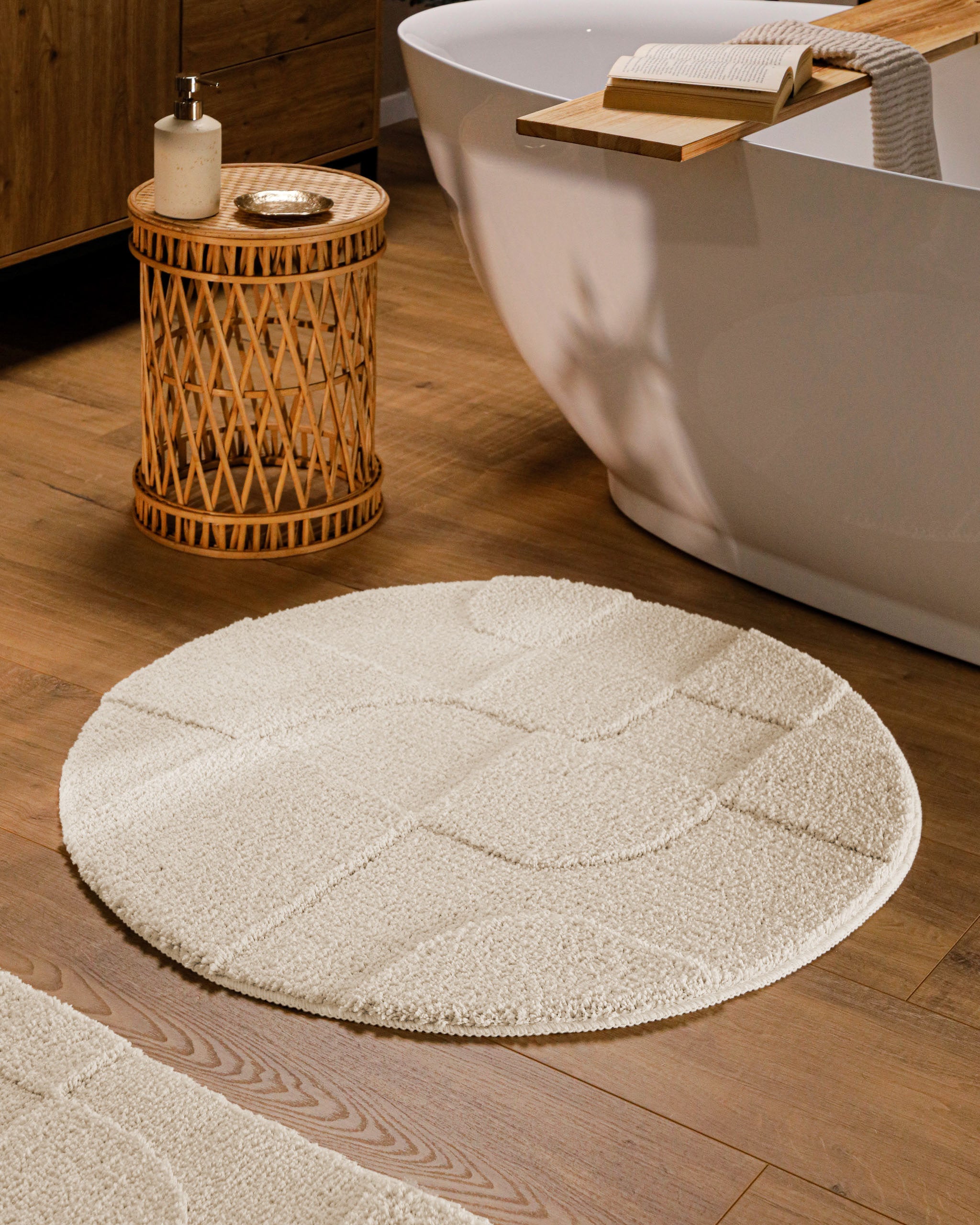 Runder Badteppich von BathKing by WECONhome Davie Creme Weiß im Badezimmer vor Badewanne auf Holzboden, strukturiertes Linienrelief im Flor sichtbar