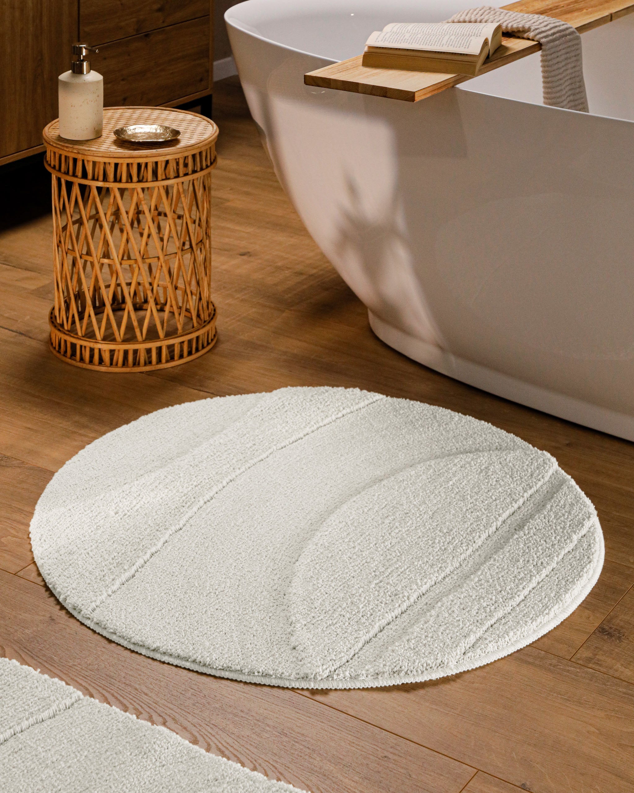 Runder Badteppich von BathKing by WECONhome Marc Weiß im Badezimmer vor Badewanne auf Holzboden, strukturiertes Linienrelief im Flor sichtbar