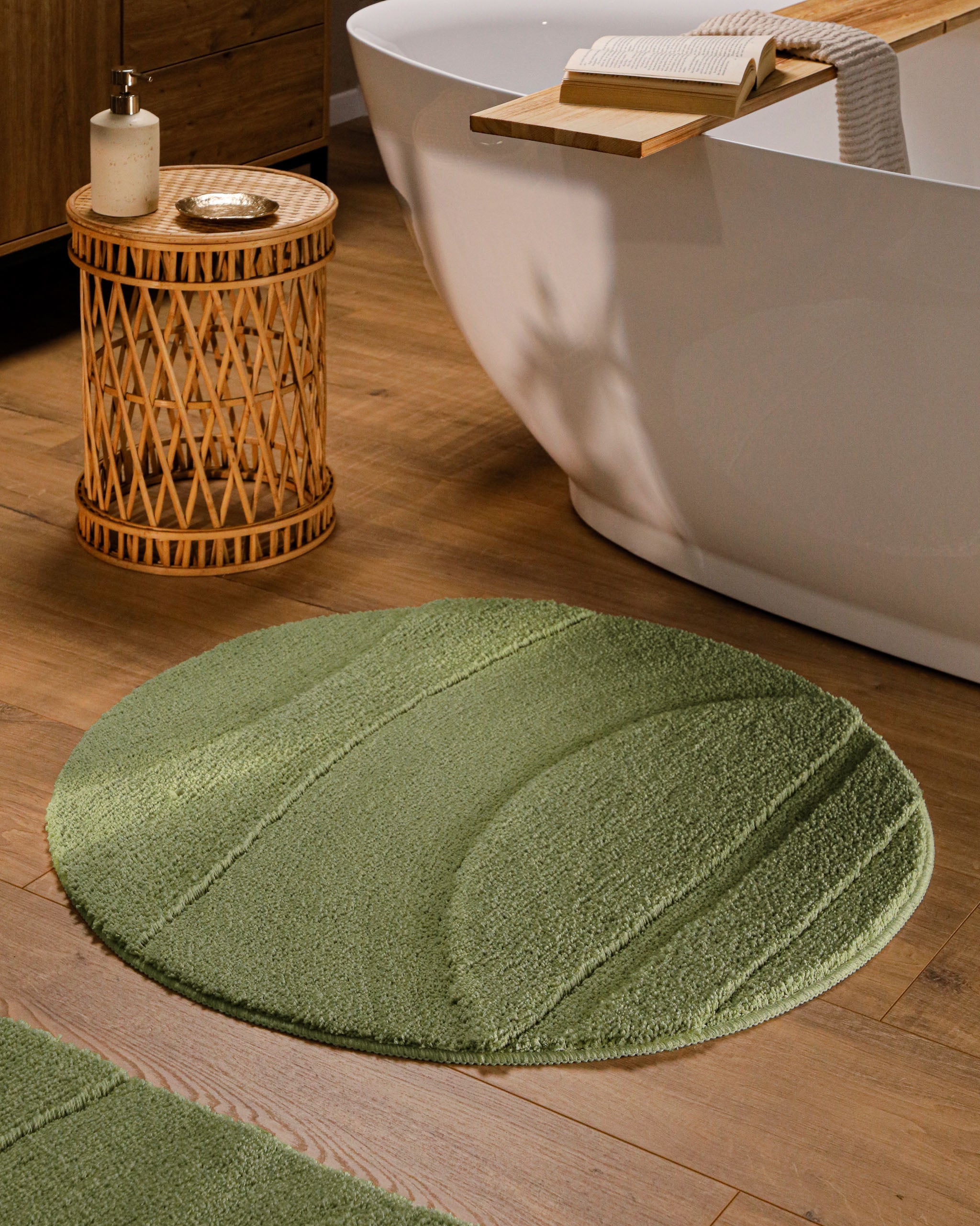 Runder Badteppich von BathKing by WECONhome Marc Reseda Grün im Badezimmer vor Badewanne auf Holzboden, strukturiertes Linienrelief im Flor sichtbar