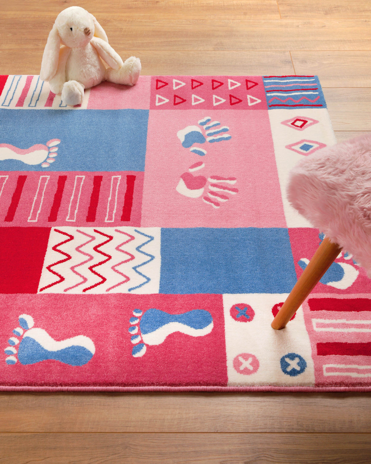 Detailaufnahme des Kinderteppichs Hand&Feet mit blauem und pinkem Fußabdruck-Motiveinem Plüschhasen  auf Holzfußboden