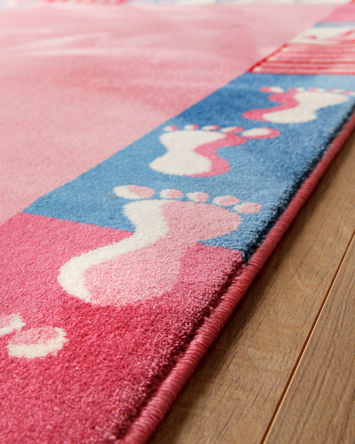 Kantendetail des Kinderteppichs Hand&Feet auf Holzboden, Rand mit blauen und pinken Fußabdrücken diagonal fotografiert