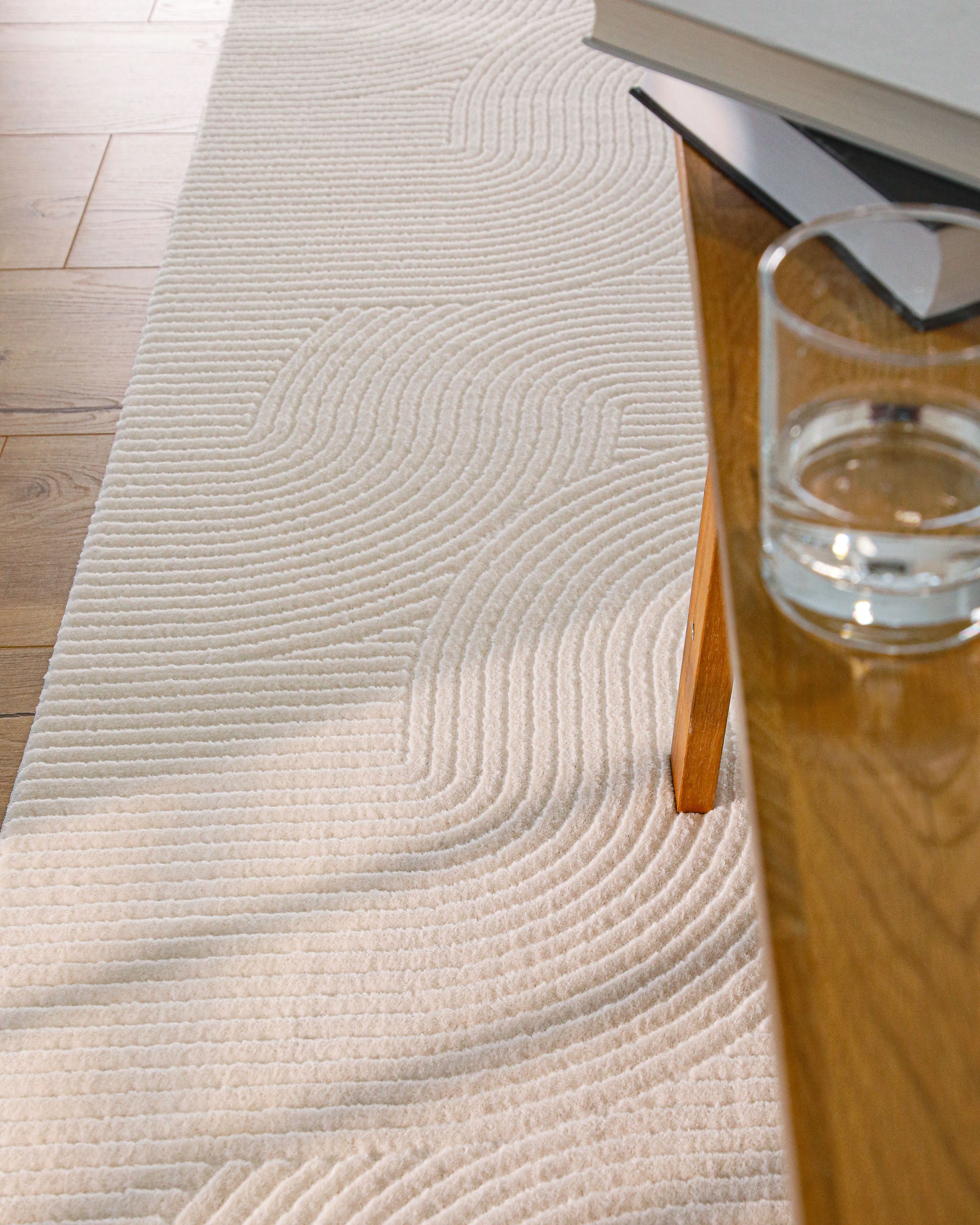 Detailansicht WECONhome Kurzflorteppich Nuuk Arc in Creme Weiß unter einem Holztisch mit einem Glas und Büchern darauf